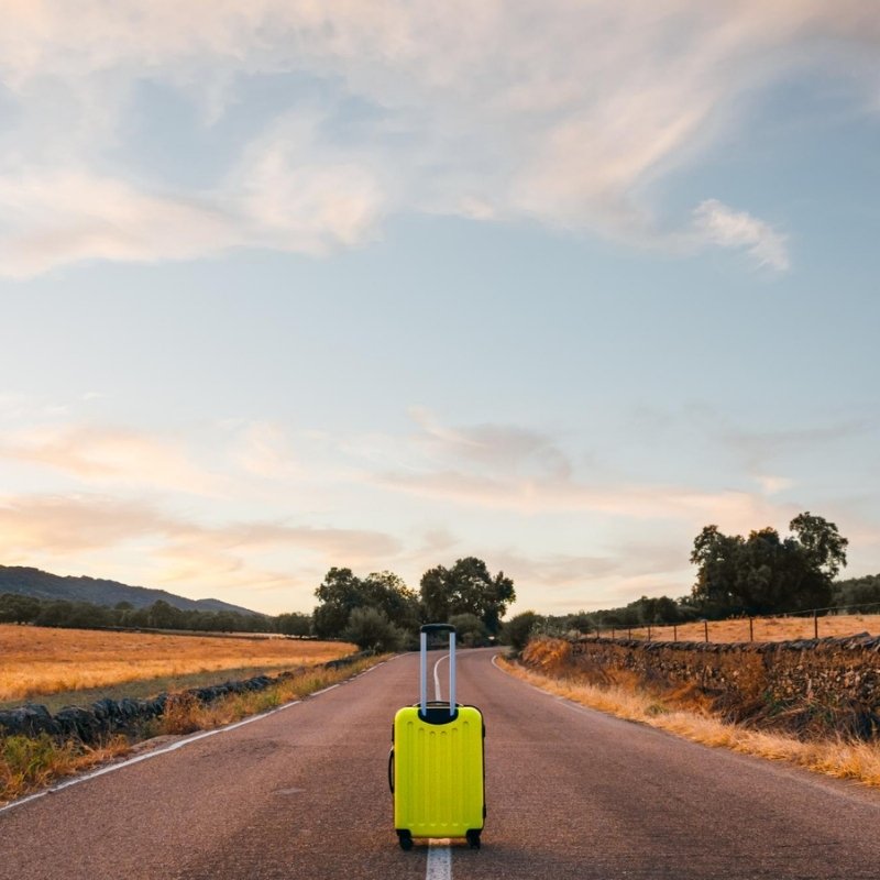 Person with yellow suitcase standing on empty rural road at sunset.